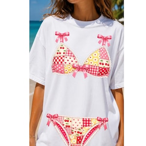 May include: A white short-sleeve t-shirt featuring a graphic print of a patchwork-style bikini set. The design includes pink bows, red cherries, yellow daisies, and red gingham patterns, with lace-like trim detailing on the bra and panty edges.