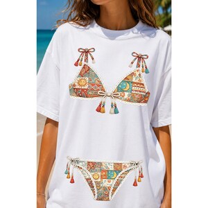 Può includere: Una t-shirt bianca a maniche corte con una stampa grafica di un bikini a due pezzi. Il bikini presenta un motivo patchwork con sole, luna e disegni geometrici nei toni dell'arancione, blu e crema, rifinito con bordi in pizzo bianco e nappe.