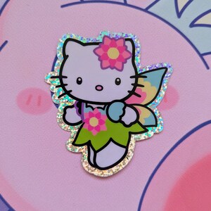 May include: A sparkly, iridescent sticker featuring Hello Kitty dressed as a fairy. She has a pink flower in her hair, green wings, and a light blue flower in her hand.