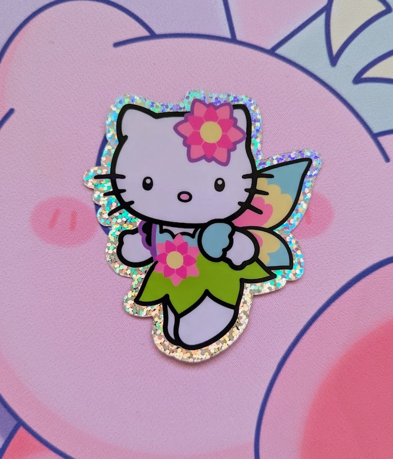 May include: A sparkly, iridescent sticker featuring Hello Kitty dressed as a fairy. She has a pink flower in her hair, green wings, and a light blue flower in her hand.