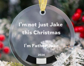 May include: A clear glass Christmas ornament with a gold ribbon hanging from a Christmas tree. The ornament has the text "I'm not just Jake this Christmas I'm Father Jake 2024" printed on it.