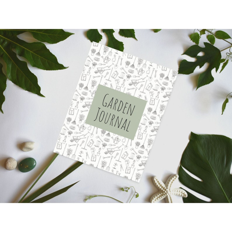 Op de afbeelding: Een wit dagboek met een groene titel die "GARDEN JOURNAL" zegt. De omslag is versierd met een patroon van zwarte en witte tuingereedschappen en planten.