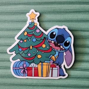 May include: A blue cartoon character, Stitch, is peeking out from behind a Christmas tree decorated with ornaments and lights. The tree is surrounded by wrapped presents with colorful ribbons.
