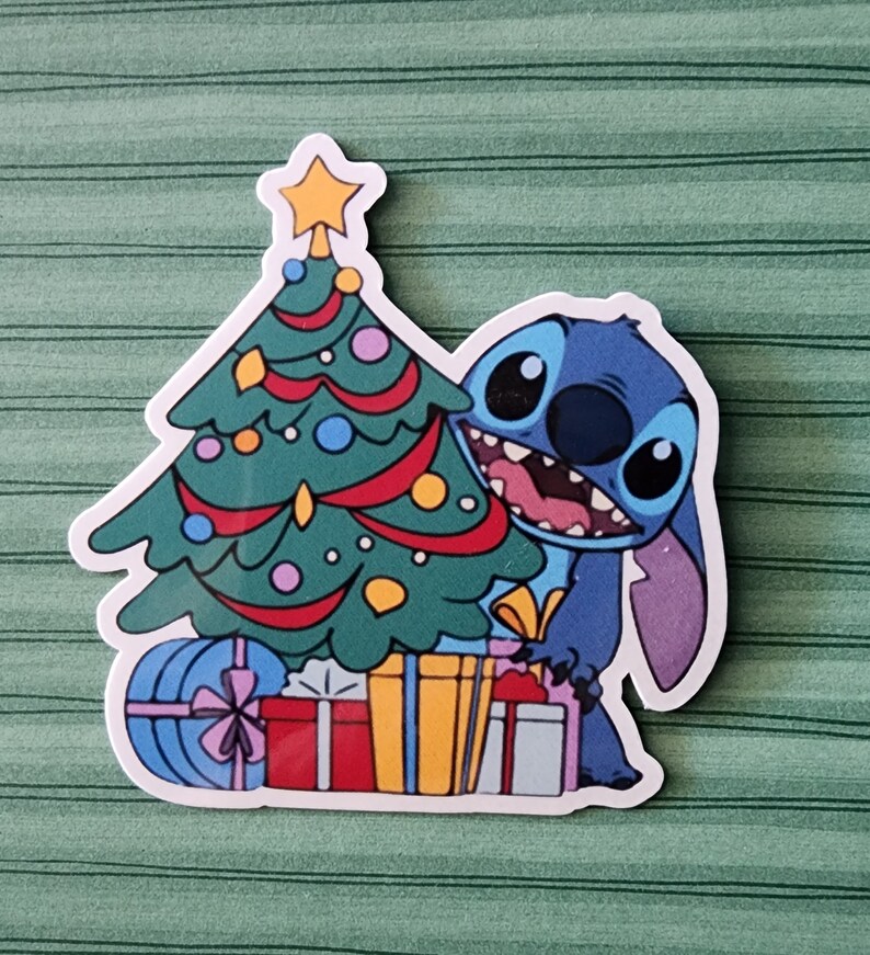 May include: A blue cartoon character, Stitch, is peeking out from behind a Christmas tree decorated with ornaments and lights. The tree is surrounded by wrapped presents with colorful ribbons.