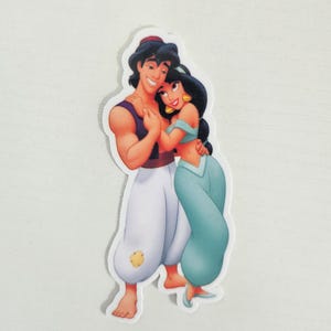 May include: A sticker featuring a cartoon depiction of Aladdin and Princess Jasmine. Aladdin is wearing a purple vest and white pants. Jasmine is in a teal outfit. They are embracing, with smiles on their faces.