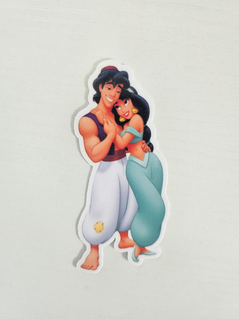 May include: A sticker featuring a cartoon depiction of Aladdin and Princess Jasmine. Aladdin is wearing a purple vest and white pants. Jasmine is in a teal outfit. They are embracing, with smiles on their faces.