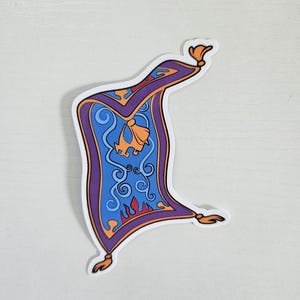 May include: A sticker featuring a stylized, cartoon-like image of a magic carpet. The carpet is primarily blue with purple borders and gold accents. The design includes swirls, a bell-shaped detail, and flame-like patterns.