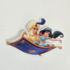 May include: A sticker featuring Aladdin and Princess Jasmine from the Disney movie, riding on a magic carpet. The carpet is blue with gold accents, and the characters are in their iconic outfits. The sticker is die-cut with a white border.