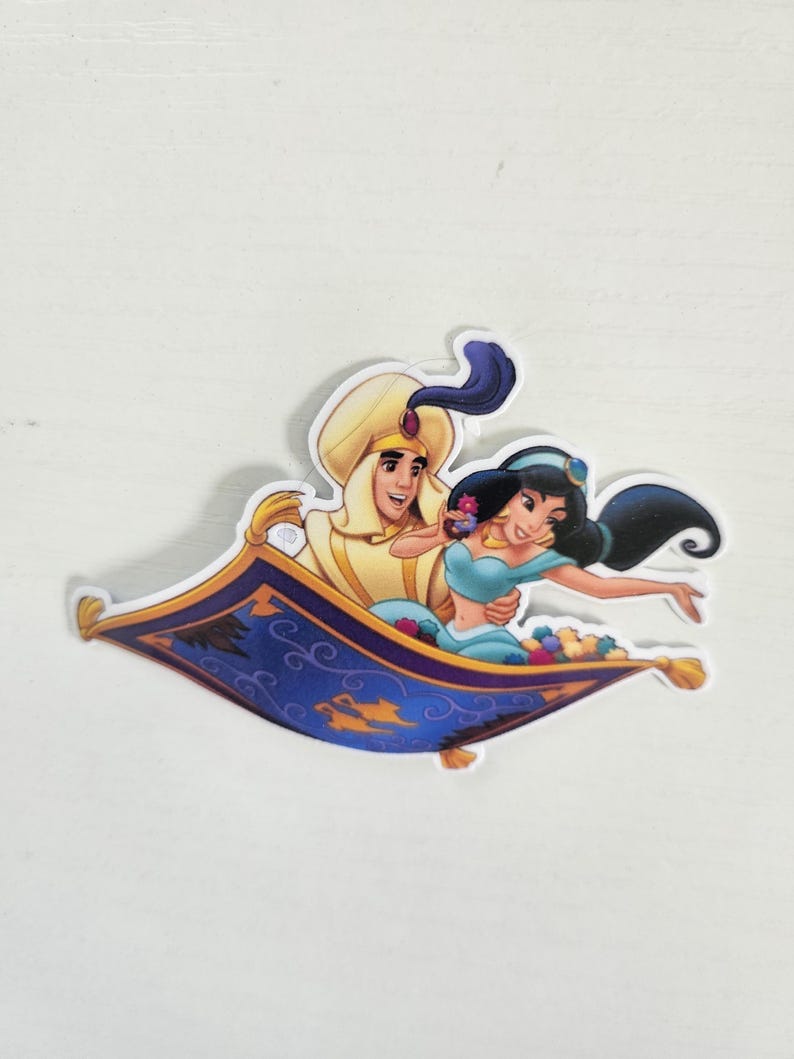 May include: A sticker featuring Aladdin and Princess Jasmine from the Disney movie, riding on a magic carpet. The carpet is blue with gold accents, and the characters are in their iconic outfits. The sticker is die-cut with a white border.