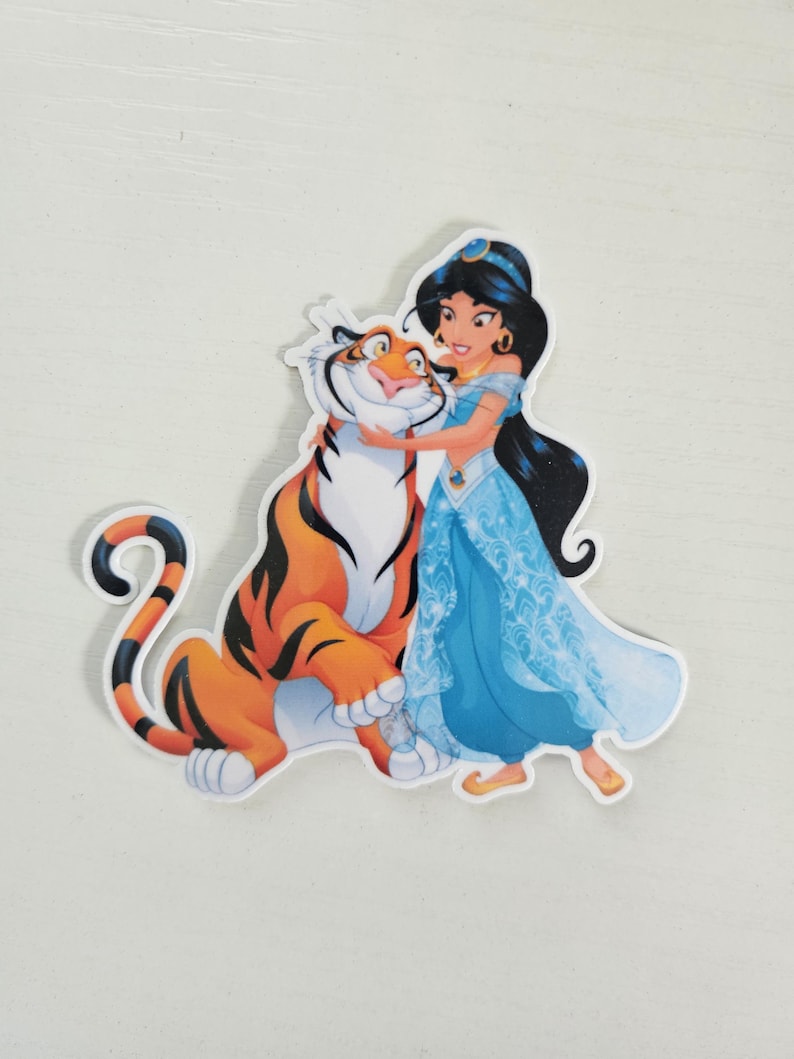 May include: A sticker featuring Princess Jasmine from Aladdin hugging Rajah the tiger. Jasmine is wearing a blue dress and has long black hair. Rajah is orange with black stripes. The sticker is on a white background.