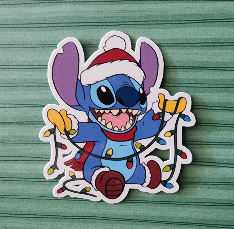 May include: A blue Stitch character wearing a red Santa hat and scarf, holding up a string of colorful Christmas lights.