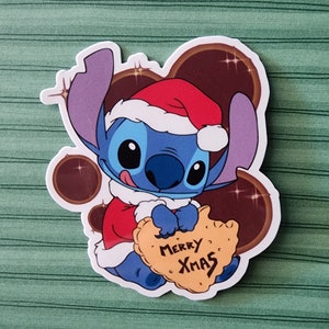 May include: A blue Stitch character wearing a red Santa hat and holding a yellow cookie that says "Merry Xmas".