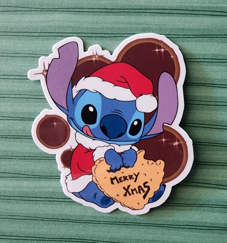 May include: A blue Stitch character wearing a red Santa hat and holding a yellow cookie that says "Merry Xmas".