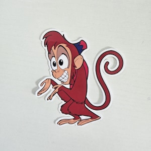 May include: A sticker featuring a cartoon monkey character in a squatting pose. The monkey is primarily red with a blue and red hat. It has a large, expressive face with a wide grin and large eyes. The sticker has a white border.