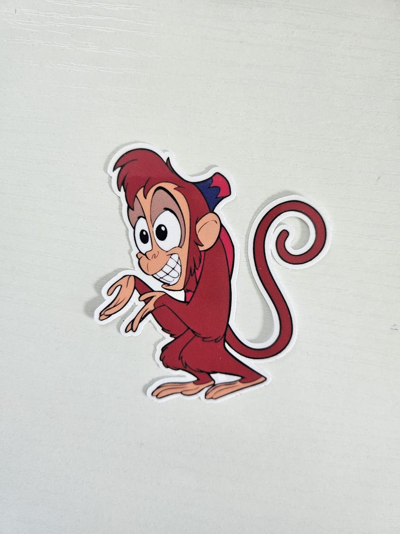 May include: A sticker featuring a cartoon monkey character in a squatting pose. The monkey is primarily red with a blue and red hat. It has a large, expressive face with a wide grin and large eyes. The sticker has a white border.