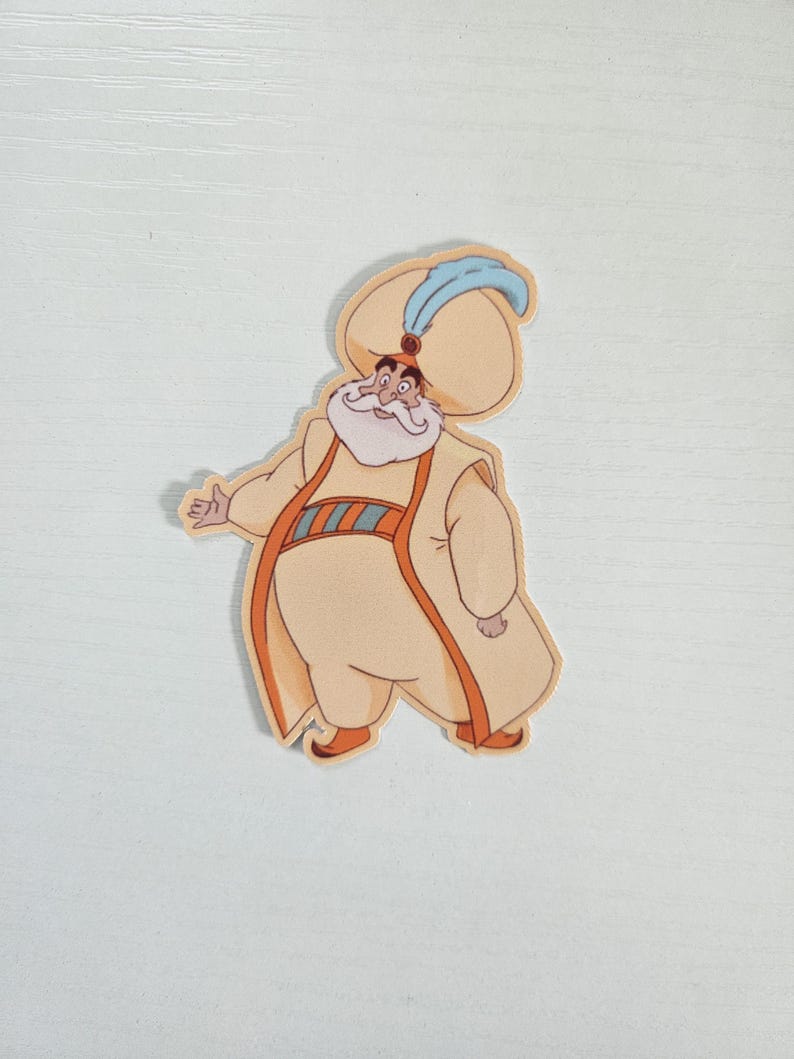 May include: A die-cut sticker featuring a cartoon character in a cream-colored robe and hat. The character has a white beard, orange accents, and a blue feather on the hat. The sticker is likely from a movie or animated series.
