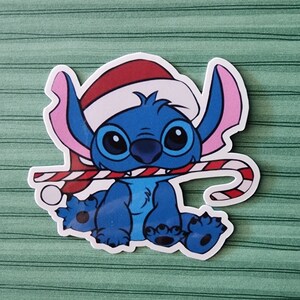 May include: A blue cartoon character wearing a red Santa hat and holding a candy cane. The character has large eyes and a wide smile.
