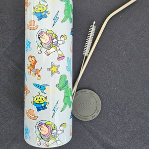 Puede incluir: Un vaso blanco con personajes de dibujos animados de Toy Story, incluyendo a Buzz Lightyear, extraterrestres y Rex. Incluye una pajita plateada, un limpiador de pajitas y una tapa gris con la palabra "coffee".