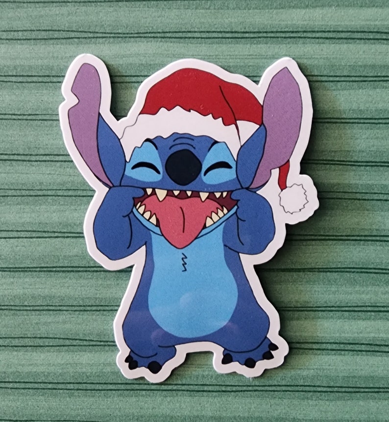 May include: A blue cartoon character with a red Santa hat, sticking out its tongue and making a silly face. The character has large ears and a wide smile.