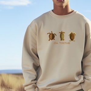 Könnte beinhalten: Cremefarbenes Sweatshirt mit drei gestickten Meeresschildkröten in Braun- und Goldtönen. Der Text "SEA TURTLES" ist unter den Schildkröten gestickt. Das Sweatshirt wird von einer Person im Freien getragen.