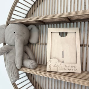 Puede incluir: Un marco de fotos de madera con una pequeña ilustración de elefante y el texto "Mein kleiner Bruder & Ich". El marco está sentado en un estante con un juguete de elefante de punto.