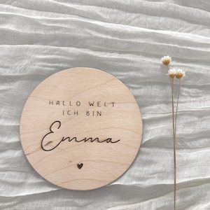 以下が含まれることがあります： 「HALLO WELT ICH BIN Emma」と小さなハートが刻まれた丸い木製のサイン。サインは明るい茶色で、自然な木目があります。背景は白く、質感のある生地です。