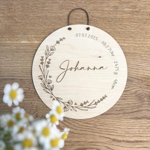 May include: A round wooden sign with the name "Johanna" in cursive script. The sign is decorated with a floral wreath and includes the date 07.07.2025, time 08:27 Uhr, weight 2475g, and height 48cm. A brown leather loop is attached for hanging.