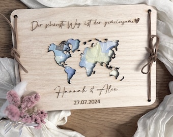 May include: A wooden guest book with a world map design and the text "Der schönste Weg ist der gemeinsame" and a heart symbol. The names "Hannah & Alex" and the date "27.07.2024" are also engraved on the book.