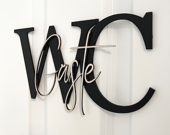 Puede incluir: Un letrero de WC de madera negra con una inscripción cursiva "Caste" en el centro. El letrero está montado en una pared blanca.