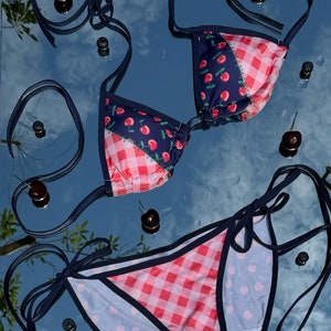Puede incluir: Un conjunto de bikini de dos piezas con un diseño lúdico. La parte superior e inferior presentan una mezcla de estampados: una tela azul marino con estampados de cerezas y un estampado de cuadros rojos y blancos. Lazos y ribetes azul oscuro completan el look.