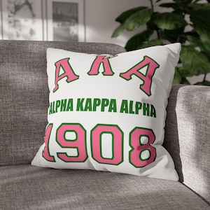 Op de afbeelding: Wit sierkussen met de tekst "ALPHA KAPPA ALPHA" in groen en het jaartal "1908" in roze. De letters "AKA" zijn ook roze. Het kussen ligt op een grijze bank.