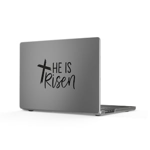 May include: A dark gray laptop featuring a black decal on the lid. The decal displays a stylized cross graphic positioned to the left of the black text that reads He Is Risen, written in a combination of block and cursive script lettering.