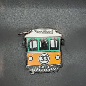 以下が含まれることがあります： 路面電車のデザインのカラフルなキーホルダー。路面電車はオレンジと緑で、白い円の中に数字の33が入っています。「SAVANNAH」の文字が上部に、「BILLY」の文字が下部に印刷されています。