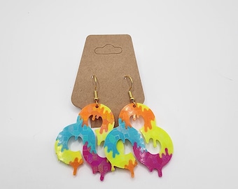 May include: Colorful, abstract earrings with a drip design. The earrings feature a neon yellow base with orange, blue, and pink accents. They hang from gold-colored hooks and are attached to a brown cardboard display card.