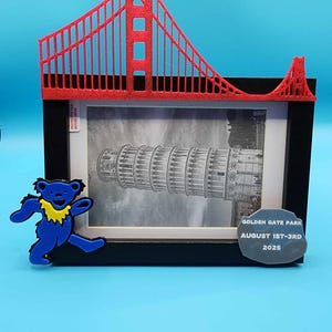 May include: A black framed photo of the Leaning Tower of Pisa, with a red Golden Gate Bridge replica on top. A blue bear-shaped pin with a yellow collar is on the left. A gray stone with text that reads "GOLDEN GATE PARK AUGUST 1ST-3RD 2025" is on the right.