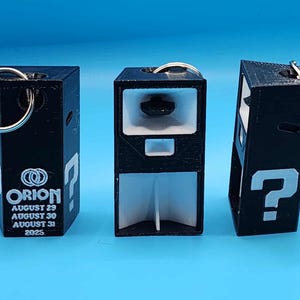 May include: Three black keychains with a rectangular design. Each keychain has a silver key ring. One keychain has the word "ORION" and dates. The other two keychains have a white question mark. The background is a solid blue.