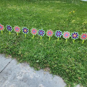 May include: A row of colorful flower decorations with pink, purple, and blue petals and yellow stems are arranged in a grassy area. The flowers are made of a plastic-like material and are likely for outdoor decoration.