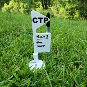 以下が含まれることがあります： 上部に「CTP」、その下に「Hole 3」と書かれた白いゴルフフラッグマーカー。円形の土台が付いており、旗の側面にある専用のスロットには黒いマーカーペンが差し込まれています。本体は白いプラスチック素材で作られています。