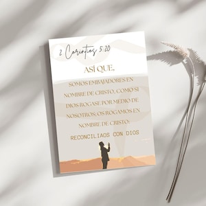 Peut inclure: Une carte rectangulaire avec le texte "2 Corintios 5:20" dans une police manuscrite. La carte présente du texte en espagnol et une illustration d'une personne avec les bras levés sur fond de coucher de soleil. La carte est sur une surface blanche.