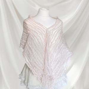 May include: A light pink, semi-sheer shawl with a crinkled, textured surface and subtle, scattered silver embellishments. The garment features a V-neckline, short sleeves, and a pointed, asymmetrical hemline with delicate fringe detailing.