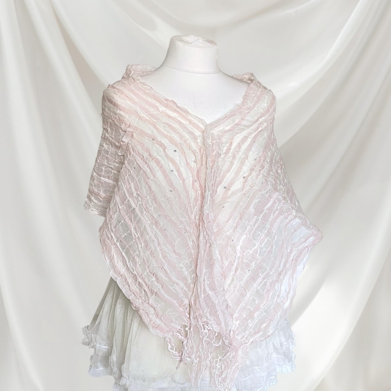 May include: A light pink, semi-sheer shawl with a crinkled, textured surface and subtle, scattered silver embellishments. The garment features a V-neckline, short sleeves, and a pointed, asymmetrical hemline with delicate fringe detailing.