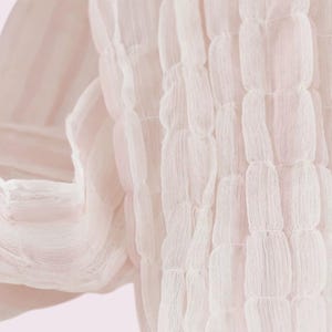 May include: A pale pink scarf featuring a textured, puckered fabric design. The material is lightweight and semi-sheer, displaying a series of vertical, gathered channels that create a repeating, soft, pillowed effect across the entire surface.