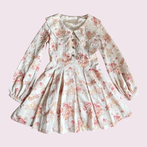 May include: A white dress with a floral pattern in pink and peach hues. The dress has a Peter Pan collar, long sleeves, and gold buttons. The skirt is pleated, and the style is vintage-inspired. The dress is suitable for a special occasion.