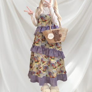 May include: A floral print dress with purple ruffles and a straw tote bag with a teddy bear decoration. The dress has a light yellow background with a floral pattern and purple ruffles. The tote bag has a teddy bear decoration.