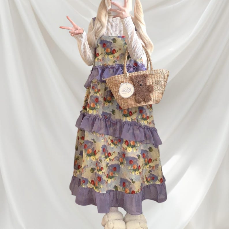May include: A floral print dress with purple ruffles and a straw tote bag with a teddy bear decoration. The dress has a light yellow background with a floral pattern and purple ruffles. The tote bag has a teddy bear decoration.