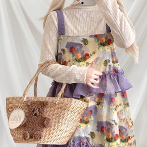 May include: A straw tote bag with a brown teddy bear applique and a tag. The bag has two handles and is held by a person wearing a floral print dress with purple accents and a cream-colored long-sleeved shirt.