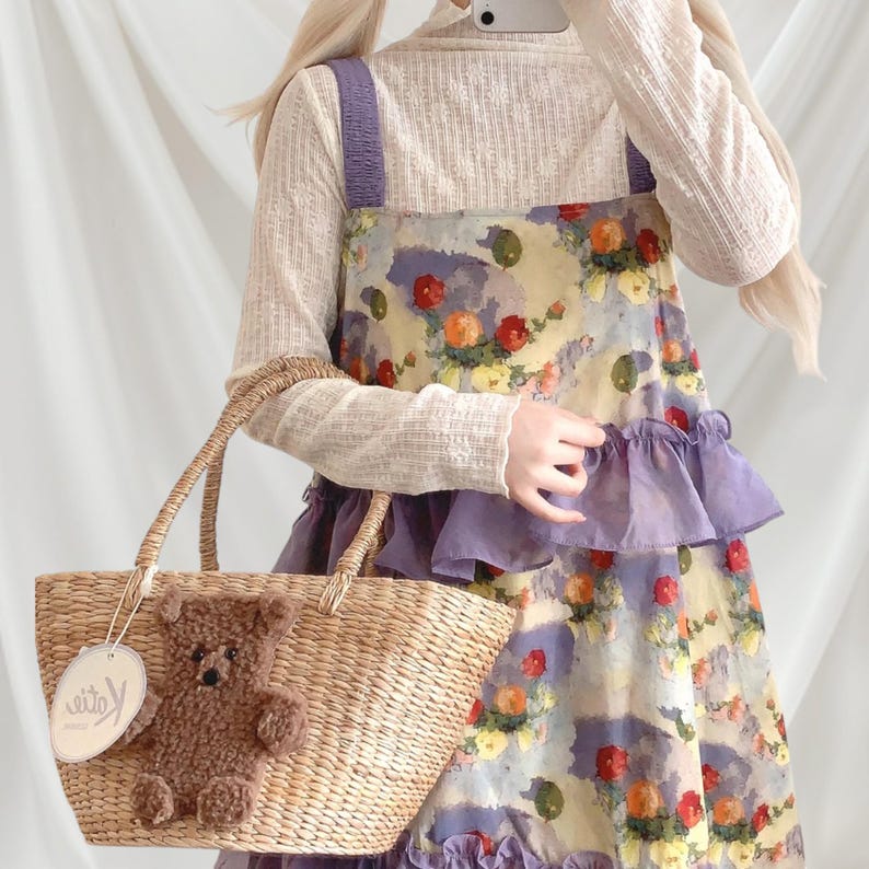 May include: A straw tote bag with a brown teddy bear applique and a tag. The bag has two handles and is held by a person wearing a floral print dress with purple accents and a cream-colored long-sleeved shirt.