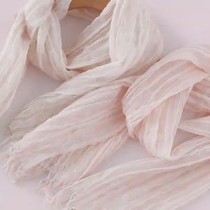 May include: A light blush pink scarf featuring a crinkled, textured fabric surface with a semi-sheer appearance. The scarf has long, loose fringe detailing at the ends and is draped in soft, flowing folds to showcase the lightweight material.