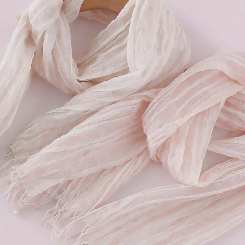 May include: A light blush pink scarf featuring a crinkled, textured fabric surface with a semi-sheer appearance. The scarf has long, loose fringe detailing at the ends and is draped in soft, flowing folds to showcase the lightweight material.