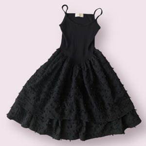 May include: A black dress with thin shoulder straps. The bodice is ribbed, and the skirt is tiered with a textured, dotted fabric. The dress has a flared silhouette, suitable for various occasions.